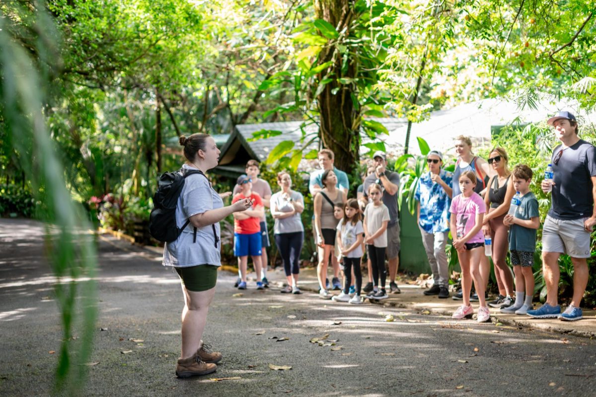Instructor with backpack speaks to a group of kids and adults outdoors in a lush, leafy park setting