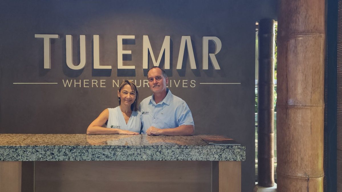 Two smiling staff members stand behind a granite reception desk in front of a dark wall with a large 'TULEMAR' sign.